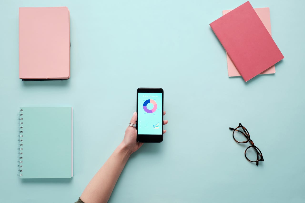 Minimalist flat lay with a smartphone displaying charts, surrounded by notebooks and glasses on a blue background.